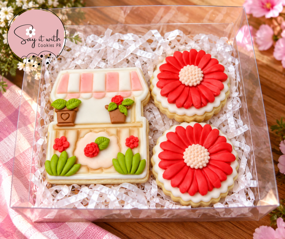 Three decorated sugar cookies in a clear plastic gift box filled with white shredded paper. One rectangular cookie is shaped like a flower shop with pink-and-white striped awning, green leaves, and red flowers.